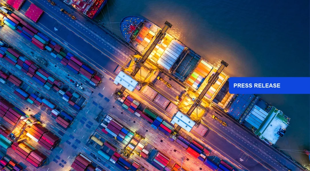 Aerial view of a busy shipping port at night, with cargo ship and containers. Press Release.
