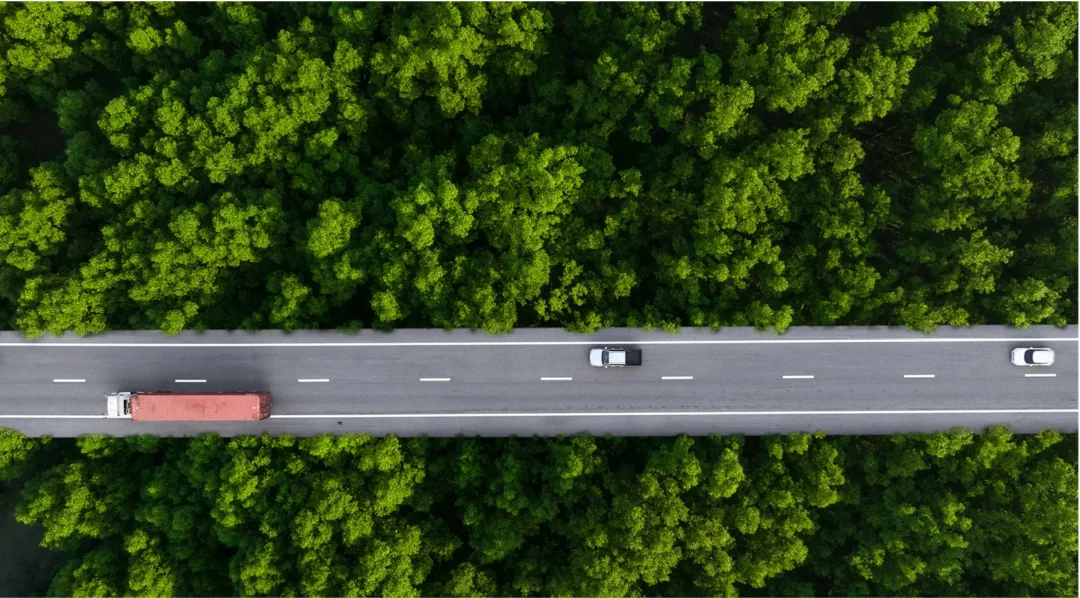 Aerial view of a highway cutting through a lush green forest, with trucks and cars on the road.