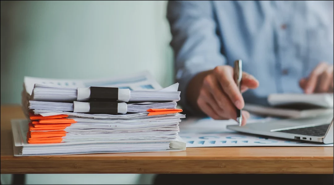 Person reviewing documents and using a laptop, related to document management.