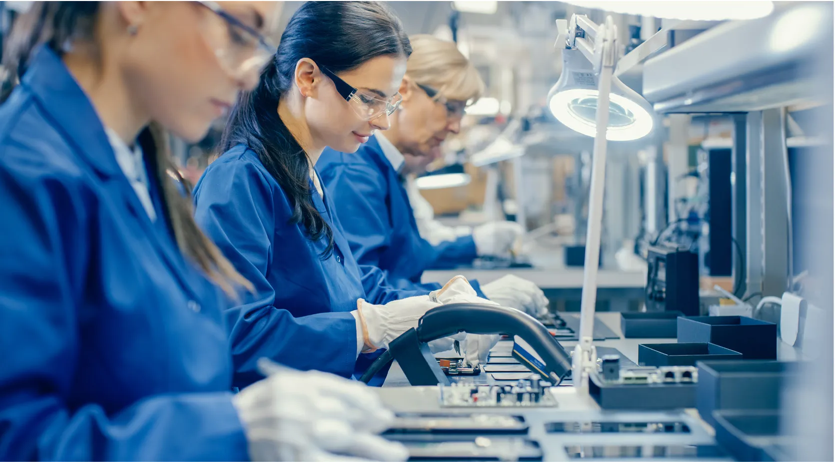 Women in blue lab coats and safety glasses assembling electronics on a production line.