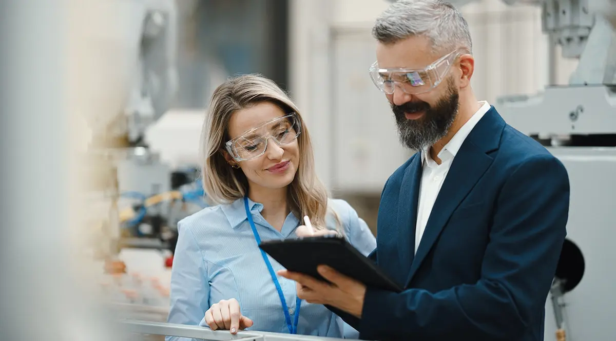 Two professionals in safety glasses review data on a tablet in a factory setting.