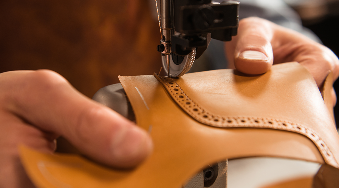 Close-up of a shoemaker using a sewing machine to craft a tan leather shoe, illustrating supply chain production.