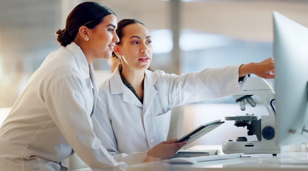 Scientists in lab coats review data on a computer and tablet next to a microscope.