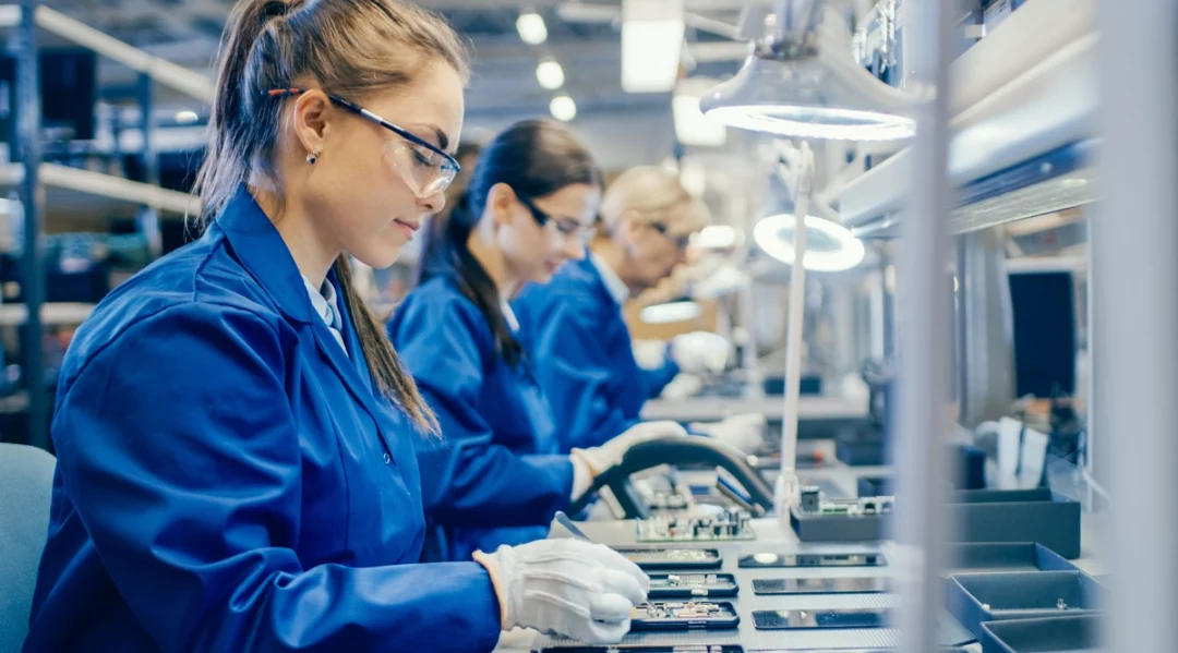 Women in blue lab coats and safety glasses assembling electronics.